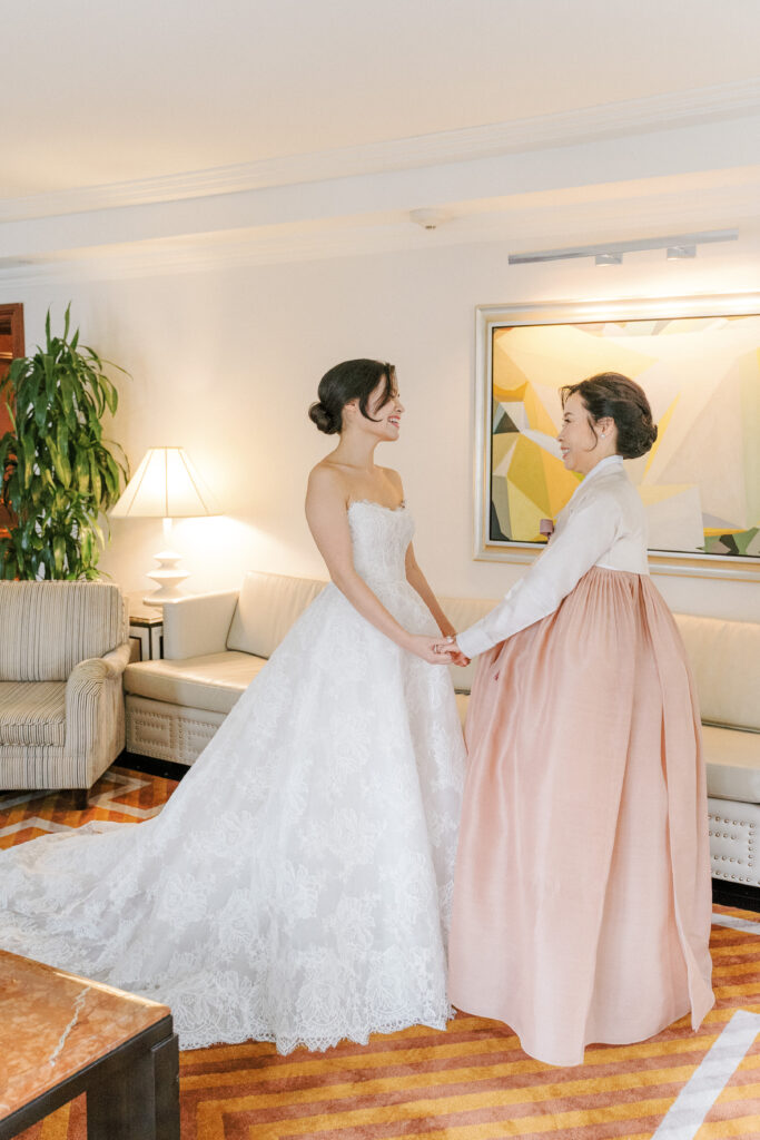 Bride in lace ballgown and mother of the bride in Korean hanbok holding hands in hotel suite at Lotte New York Palace Hotel