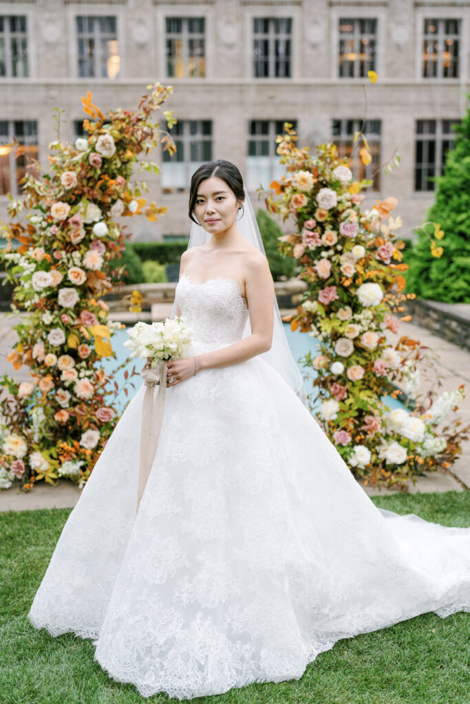 Asian bride with natural makeup in lace ballgown standing in front of fall wedding arch with pink and white roses and orange fall foliage 