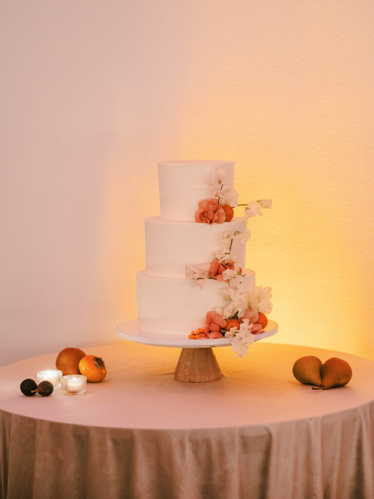 Simple 3-tier buttercream wedding cake on wood stand with orange and blush flowers and fall fruit on table with velvet linen 