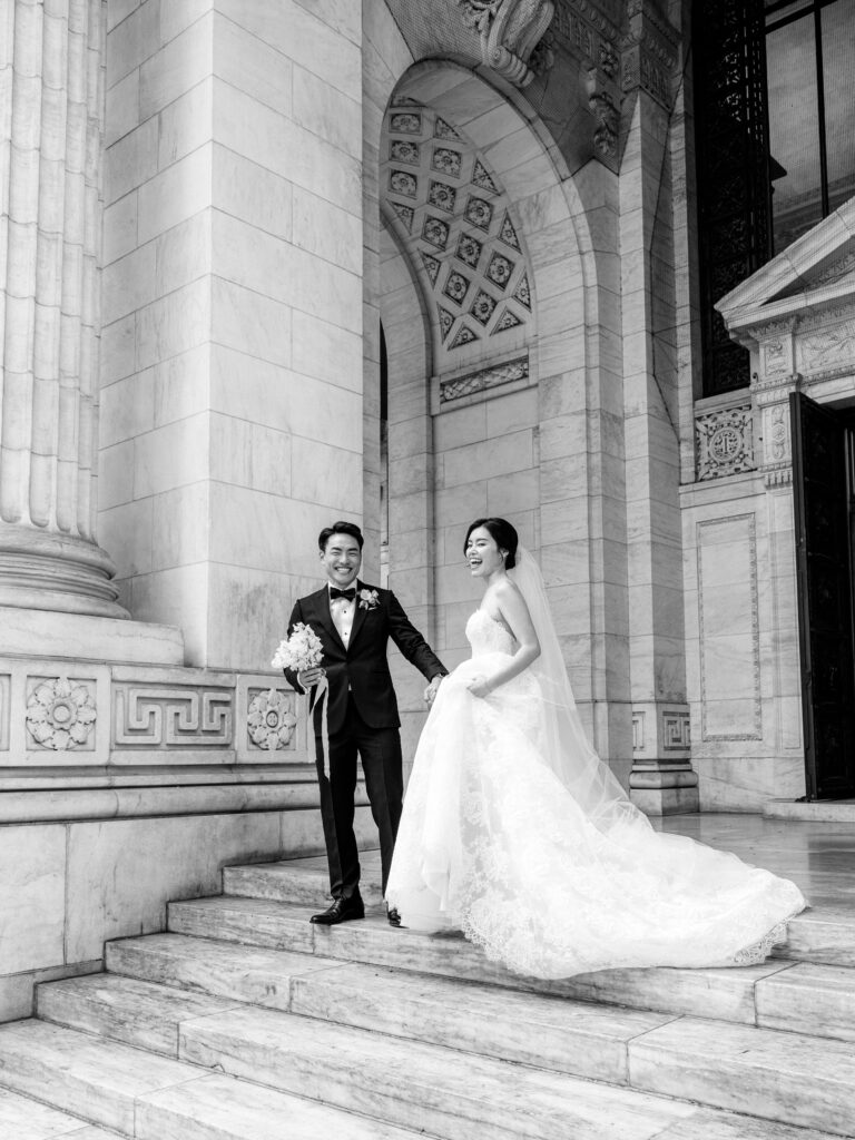 Bride and groom laughing on front steps of New York Public Library in black and white on wedding day