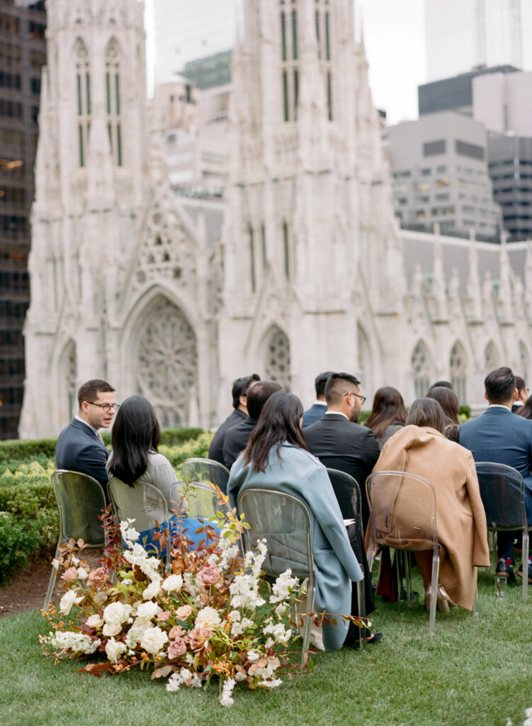 Wedding guests sitting in ghost chairs with growing garden florals at back of aisle for wedding ceremony overlooking St. Patrick's Cathedral 