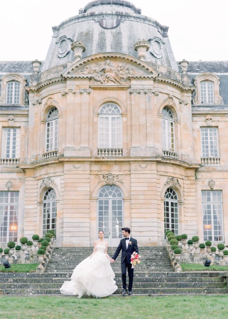 bride and groom walking outside Château de Champlâtreux holding hands and wearing ballgown and tuxedo