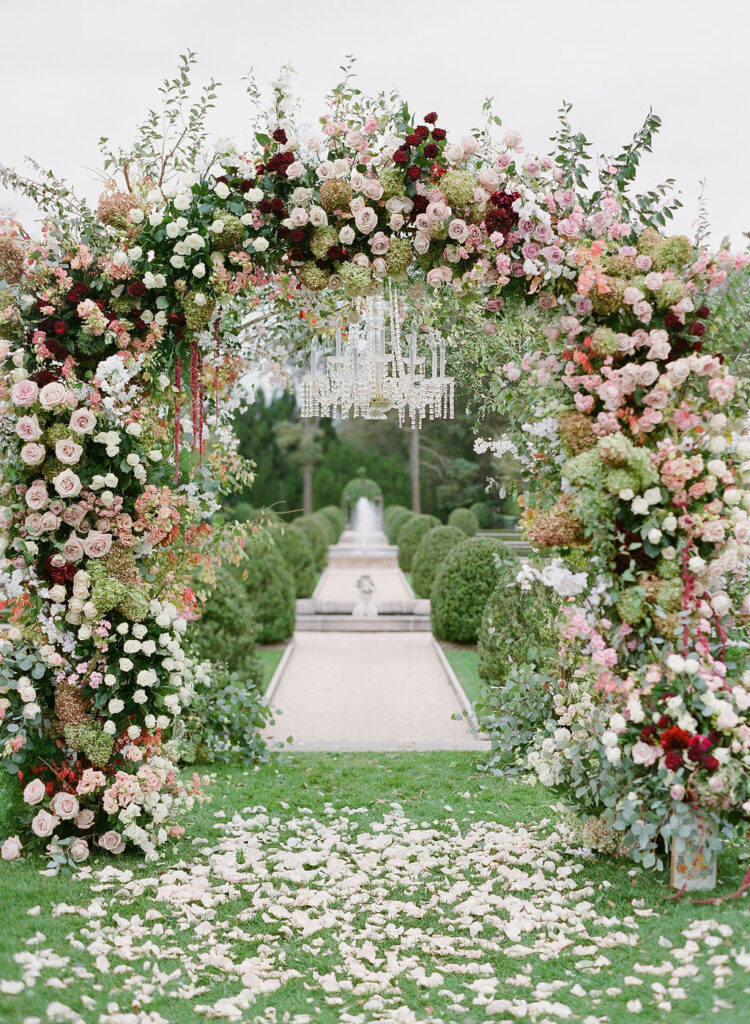 blush, red, and burgundy lush floral arch with chandelier and rose petals overlooking oheka castle gardens in new york luxury wedding