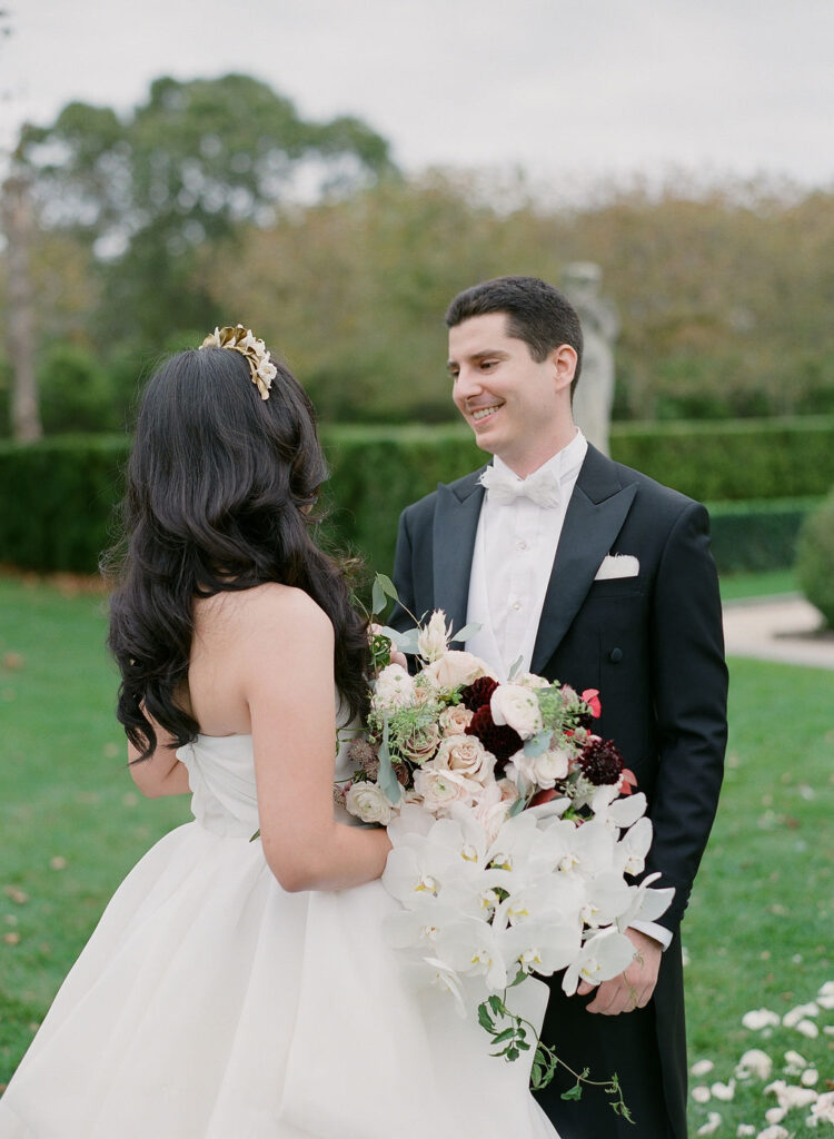 groom in tuxedo smiling at bride with long wavy dark hair in down style holding orchid bouquet on wedding day at oheka castle 