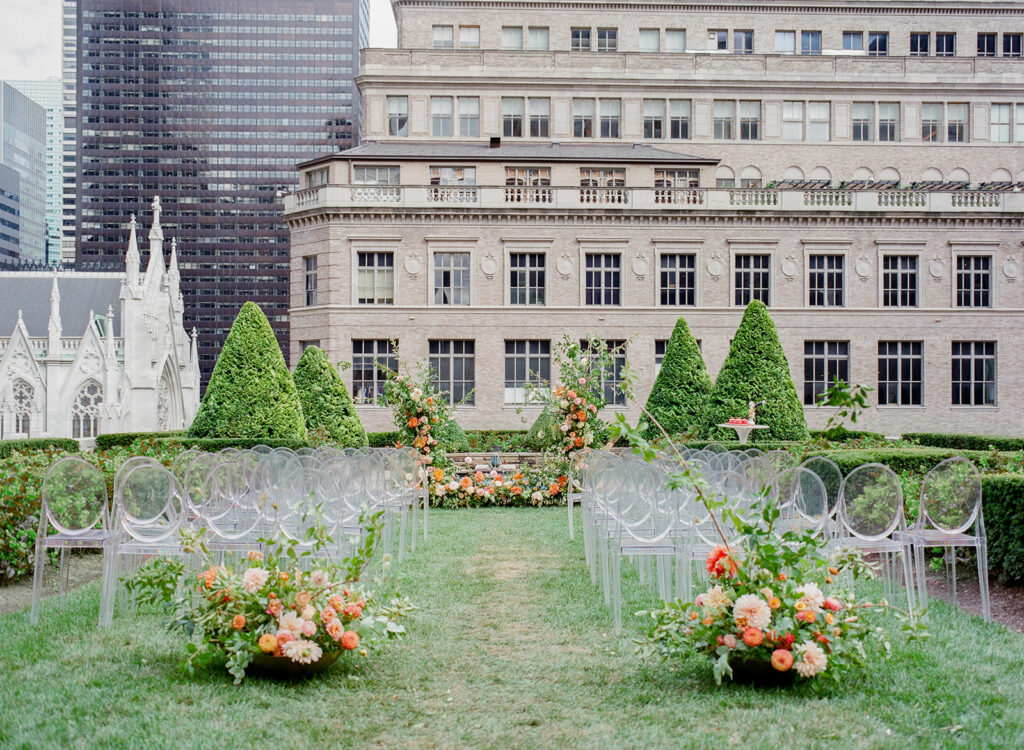 wedding ceremony in rooftop garden with ghost chairs and orange and coral florals at end of aisle 